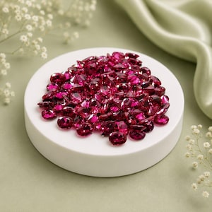 May include: A collection of numerous small, round, faceted gemstones in a deep, vibrant magenta-red colour. These loose, brilliant-cut stones are piled together in a cluster on top of a smooth, circular, white display platform.