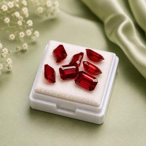 May include: A collection of six faceted, deep red gemstones arranged on a white square display pad. The stones feature emerald-cut and rectangular shapes with sharp, reflective edges, showcasing a vibrant crimson hue and high clarity throughout.
