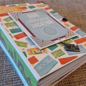 May include: A travel-themed photo album with a light blue cover featuring the text "Our Amazing Vacation." The album is decorated with suitcase and travel-related graphics in various colors. A small camera charm hangs from the album.