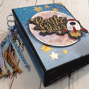 May include: A rectangular scrapbook with a dark blue space-themed cover featuring gold stars, a circular moon cutout, and a rocket ship cutout. The text "Reach for the stars" is written in gold script. A multi-textured tassel with charms hangs from the side.