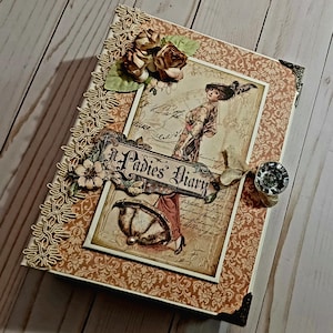 May include: A rectangular diary with a tan and brown damask patterned cover. The front features a central illustration of a person with an umbrella, the text A Ladies Diary, decorative fabric roses, gold trim, and a clear crystal-like button.