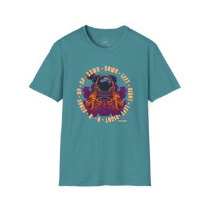 Könnte beinhalten: Türkisfarbenes T-Shirt mit einer Grafik eines Astronauten, der ein Videospiel spielt. Das Design enthält die Wörter "UP, DOWN, LEFT, RIGHT, START, B, A" in einem kreisförmigen Muster um den Astronauten. Der Astronaut trägt einen orangefarbenen und lila Raumanzug.
