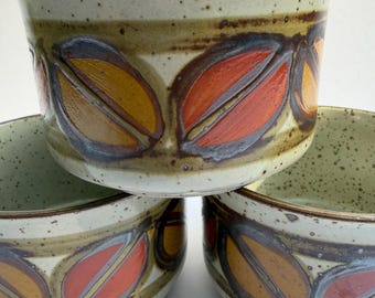 May include: A set of three ceramic bowls featuring a speckled off-white base with a horizontal band of stylized leaf motifs in shades of orange, yellow, and brown. The bowls have a wide, open design with a smooth, glazed finish and dark brown rims.