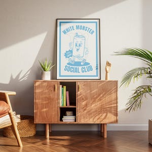 May include: A framed poster with a light blue and white design featuring a cartoon can character with the text "WHITE MONSTER SOCIAL CLUB". The poster is displayed on a wooden cabinet with a plant and decorative items.