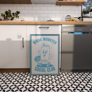May include: A framed poster with a light blue border and the words "WHITE MONSTER SOCIAL CLUB" in a retro font. The poster features a cartoon can character with a smiling face and the words "STILL AWAKE". The poster is displayed in a kitchen.