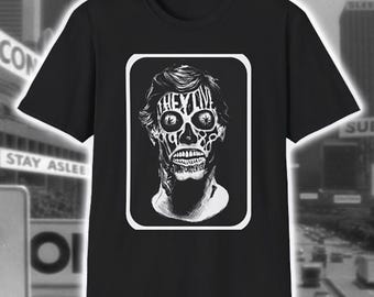 May include: Black t-shirt featuring a white and black graphic of a skull-like face with the word "THEY LIVE" in the forehead area. The design is framed in a white rectangle. The shirt is a crew neck style.