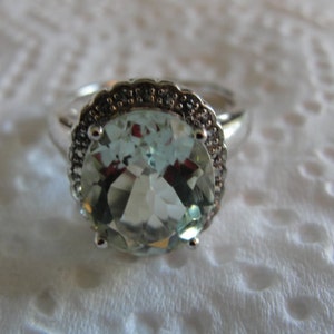 Könnte beinhalten: Ein silberner Ring mit einem großen, oval geschliffenen, hellgrünen Edelstein. Der Edelstein ist von einem Kranz kleinerer, dunkler Steine umgeben. Der Ring hat ein schlichtes, poliertes Silberband. Der Ring liegt vor einem weißen, strukturierten Hintergrund.