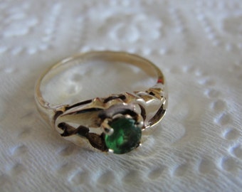 May include: A gold-colored ring with an emerald-green gemstone. The ring has an ornate design around the gemstone, with decorative cutouts on the band. The gemstone is round and set in a prong setting. The ring is on a white surface.