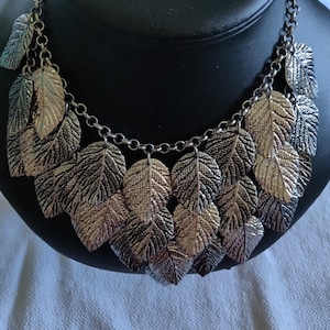 May include: A silver necklace with a chain and multiple leaf-shaped pendants. The leaves are layered and have a textured surface.