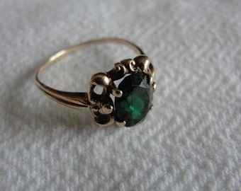 May include: A gold ring featuring a large, oval-cut, dark green gemstone. The ring has an ornate, filigree-style setting around the stone and a slender band. The ring is displayed on a white, textured surface.