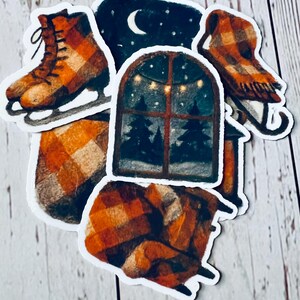 May include: A collection of winter-themed stickers featuring a pair of ice skates, a sled, a window with a snowy scene, and cozy blankets. The stickers have a warm color palette of orange, brown, and white.