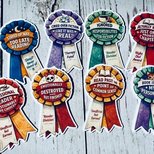 May include: A collection of colorful, ribbon-shaped stickers with humorous text about reading. Designs include phrases like "Stayed Up Way Too Late Reading" and "Cried Over Fiction Like It Was Real." Each sticker has a unique color scheme and design.