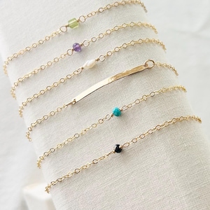 May include: A collection of delicate gold chain bracelets. Each bracelet features a unique charm, including a green gemstone, a purple gemstone, a pearl, a gold bar, a turquoise bead, and a black bead. The bracelets are displayed on a white cloth.