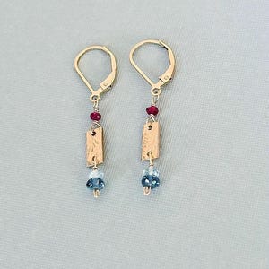 May include: A pair of gold leverback earrings featuring a small red bead, a rectangular gold charm with a textured surface, and a faceted blue bead at the bottom. The earrings are constructed with gold wire links connecting each component vertically.