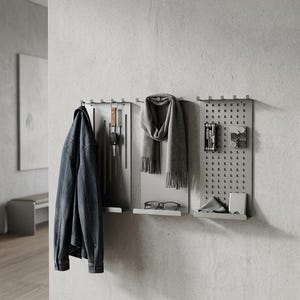May include: A contemporary grey wall organiser with three panels. A denim jacket hangs on the left, a grey scarf and glasses in the middle, and tools and accessories on the right. The system is mounted on a concrete wall.