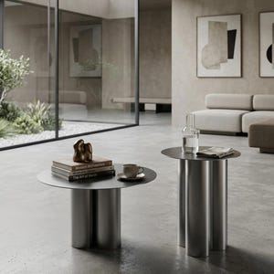 Peut inclure: Deux tables d'appoint rondes en métal avec des bases cylindriques. La table la plus basse porte une pile de trois livres intitulés Modern Architecture, Minimalist Design et The Art of Metal, surmontée d'une sculpture dorée. La table haute porte une carafe en verre et une tasse.