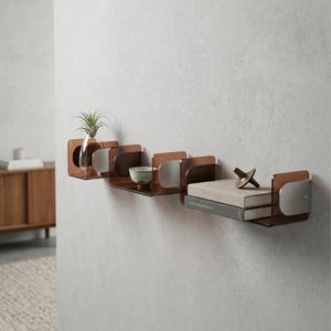 Puede incluir: Una serie de estanterías modernas montadas en la pared, en un acabado marrón cálido y plateado. Cada estante contiene objetos decorativos, incluyendo una pequeña planta en un jarrón de cristal, un cuenco de cerámica, libros y una escultura de latón. Las estanterías están montadas en una pared gris claro.