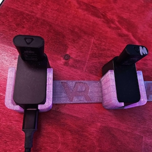 May include: Two black VR controllers with charging cables are mounted in a white plastic holder with the word "VR" printed on it.