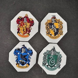 May include: Four white octagonal fabric patches featuring the Hogwarts house crests. The crests are Gryffindor, Hufflepuff, Ravenclaw, and Slytherin. Each crest is surrounded by a white border and features the house colours and mascot.