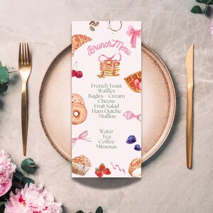 May include: A watercolour brunch menu card featuring illustrations of food items, including waffles, pancakes, and fruit. The menu lists items such as French toast, bagels, and mimosas. The card is on a plate with gold cutlery, with pink flowers in the background.