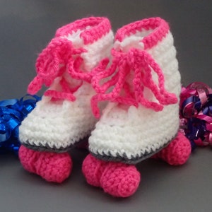 May include: White crocheted baby booties shaped like roller skates with pink laces and wheels. The booties are on a gray surface with blue and pink ribbon.