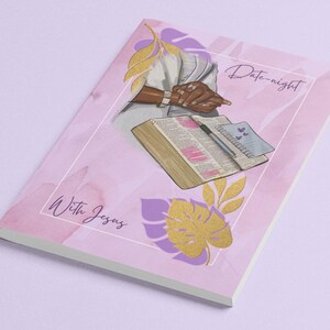 May include: A pink and purple notebook with a watercolour background. The notebook features a drawing of a person holding a Bible open to a page with hearts drawn on it. The text "Date-night with Jesus" is written on the cover.