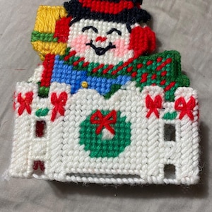 May include: A handmade snowman decoration with a black hat, red earmuffs, and a green and red scarf. The snowman is smiling and holding a yellow broom. The decoration is attached to a white fence with red bows and a green wreath.