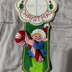 Könnte beinhalten: Ein Weihnachtsornament aus Filz mit einem lächelnden Elf, der eine Zuckerstange hält. Das Ornament hat ein grünes Banner mit goldenem Rand und eine weiße, geschwungene Oberseite mit den roten Worten "MERRY CHRISTMAS". Grüne Stechpalmen und rote Pailletten schmücken das Ornament.