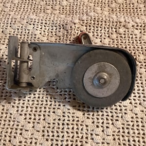 May include: A vintage metal tool with a grinding wheel and a red handle. The tool is silver and has a rectangular body with a circular grinding wheel on one end. The background is a cream-colored crocheted surface.