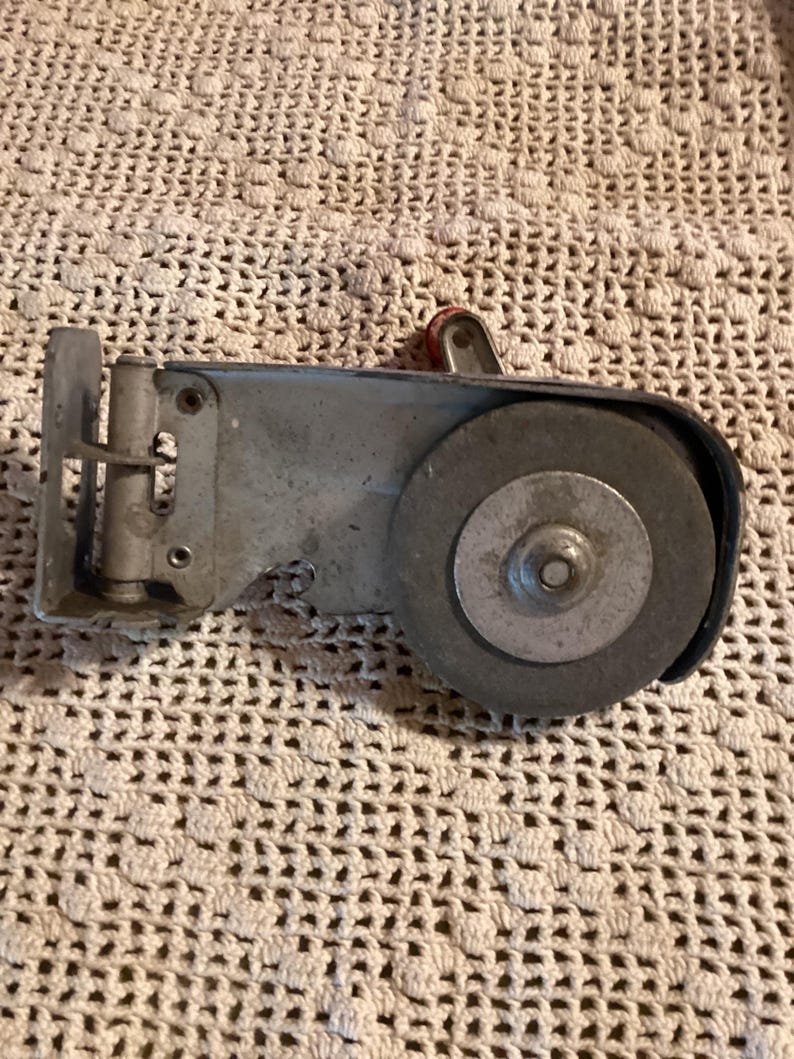 May include: A vintage metal tool with a grinding wheel and a red handle. The tool is silver and has a rectangular body with a circular grinding wheel on one end. The background is a cream-colored crocheted surface.