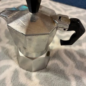 May include: A silver-colored aluminum moka pot with an octagonal base and upper chamber. It features a black plastic handle and a matching black octagonal knob on the hinged lid. The metal surface shows a brushed texture with visible signs of use.