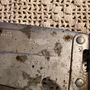 May include: Close-up of a vintage Swing-A-Way can opener. The metal opener shows signs of wear, with rust spots and faded text. The text includes "Swing-A-Way" and "St. Louis, MO, USA". The opener has a purple handle.