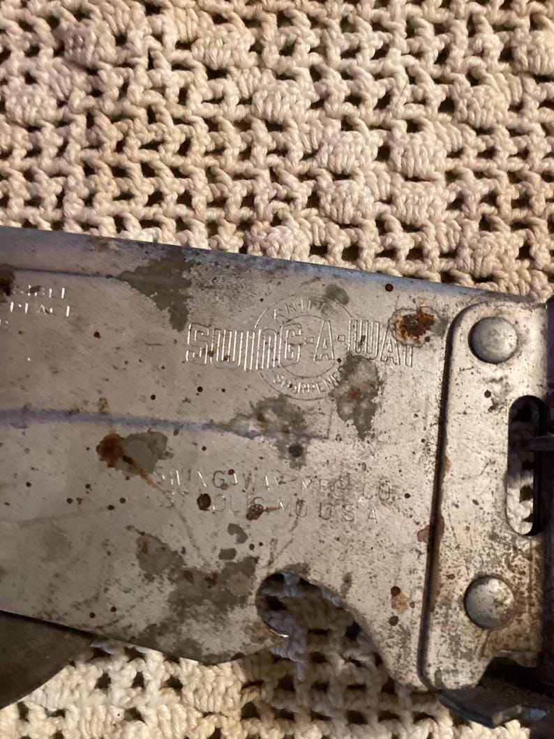 May include: Close-up of a vintage Swing-A-Way can opener. The metal opener shows signs of wear, with rust spots and faded text. The text includes "Swing-A-Way" and "St. Louis, MO, USA". The opener has a purple handle.