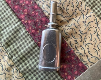 May include: A silver oil can with a long spout and a circular indentation on the front. The can is sitting on a patterned fabric.