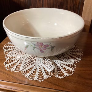 May include: A large, off-white ceramic bowl with a floral design featuring pink and purple flowers. The bowl sits on a cream-colored, crocheted doily. The rim of the bowl has a dark, distressed edge. The bowl is likely used for serving or mixing.