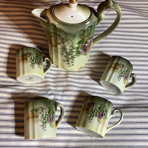 May include: A vintage tea set featuring a teapot and four matching mugs. The set is painted with a green and white color scheme, with a floral design of grapes. The teapot has a gold-colored knob on the lid.
