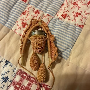 May include: A vintage sewing thimble holder featuring a metallic thimble encased in a crocheted orange cover. The holder is suspended from a curved, bone-colored frame wrapped in matching orange thread, with two small crocheted orange weights attached.