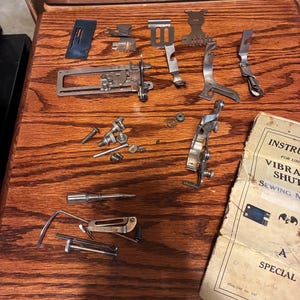 May include: A collection of vintage sewing machine parts, including various metal pieces, screws, and a manual. The parts are arranged on a wooden surface, showcasing their intricate designs and aged appearance. The manual is titled "Instructions for Use Vibrating Shuttle Sewing Machine."