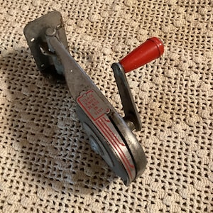 May include: Vintage metal knife sharpener with a red handle. The sharpener has a silver body with red accents and the text "PLACE KNIFE BLADE IN SLOT". The sharpener is designed to sharpen knives.