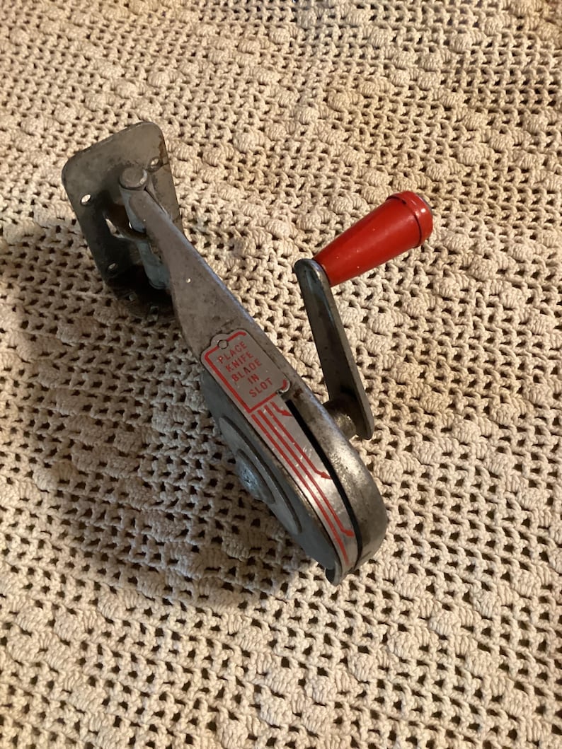 May include: Vintage metal knife sharpener with a red handle. The sharpener has a silver body with red accents and the text "PLACE KNIFE BLADE IN SLOT". The sharpener is designed to sharpen knives.