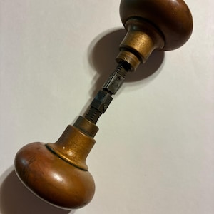 May include: A pair of round, smooth, brown wooden door knobs connected by a central metal spindle. The knobs feature a warm, polished finish with visible wood grain, attached to brass-toned metal bases that taper into a threaded square rod.