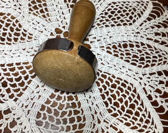 May include: A wooden meat tenderizer with a rounded head and a metal band, resting on a white, crocheted doily. The handle is a light brown color, and the doily has a detailed, openwork pattern.