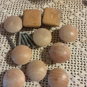 May include: A collection of unfinished wooden knobs and square pulls, along with several silver screws. The knobs are round and the pulls are square. The items are arranged on a cream-colored crocheted surface.