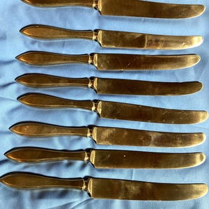 May include: Seven vintage butter knives with gold-colored handles and blades. The knives are arranged in parallel on a blue surface. The blades have a slightly curved shape, and the handles have a rounded, elongated design. The knives are likely made of stainless steel.