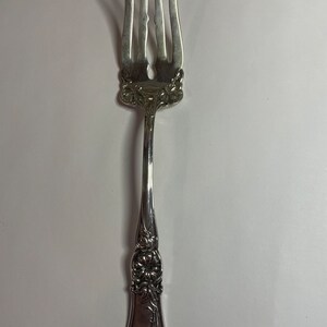May include: Silverware fork with four tines and ornate floral detailing. The handle features embossed flowers and leaves. The fork is made of a shiny, reflective metal, likely silver or a silver-colored alloy. The tines are slightly curved.