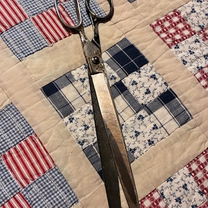 May include: A pair of vintage metal sewing shears with two oval finger loops and long, pointed blades. The metal shows signs of age, including surface wear and discoloration, with a central pivot screw connecting the two blades of the scissors.