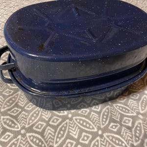 May include: A set of two oval-shaped, navy blue enamel roasting pans with white speckles. The pans have handles and lids, perfect for baking and cooking. The pans are stacked on top of each other.