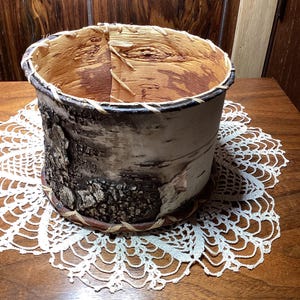 May include: A handcrafted birch bark basket with a natural, rustic appearance. The cylindrical container features a textured exterior with dark brown and light beige tones. The rim is reinforced with woven material. The basket sits on a white lace doily.