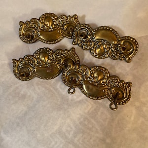 May include: Three ornate brass drawer pulls with a decorative scroll design and a loop on each side. The pulls are in a vintage style and have a worn, antique finish.