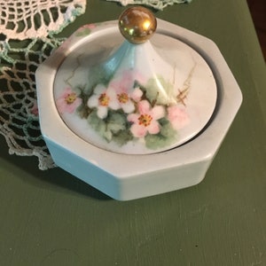 May include: A white, hexagonal trinket box with a lid featuring a floral design. The lid has a gold-colored knob. The flowers are pink and white with green leaves. The box is on a green surface.
